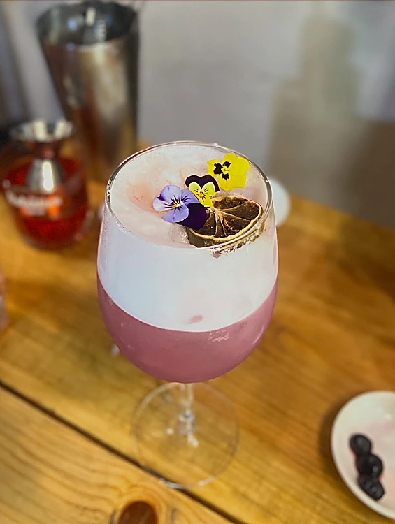 Lavanda Sour en copa, color lila con espuma blanca, flores comestibles y cítrico deshidratado