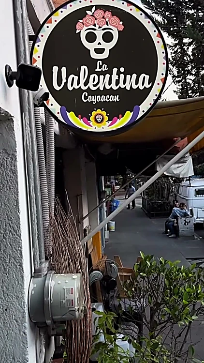Letrero redondo de La Valentina Coyoacán con calavera y flores, colgado sobre la banqueta